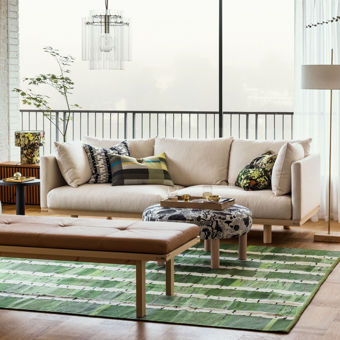 A bright living room with a beige STOCKHOLM 2025 3-seat sofa by a bench and pouffe sitting on a green, black, and white rug.