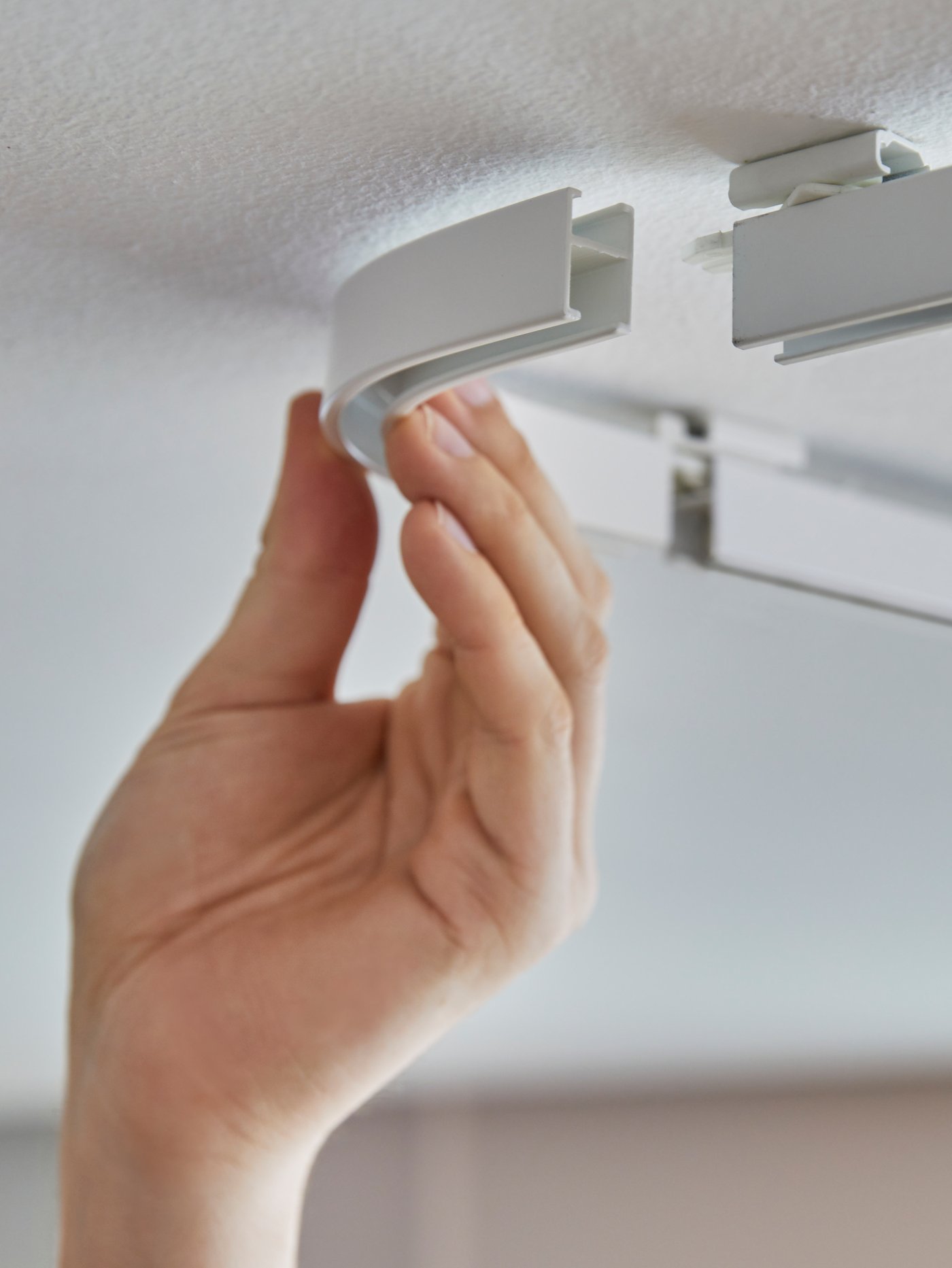 La main d’une personne qui raccorde un élément d’angle de rail pour rideaux VIDGA à des rails simples VIDGA fixés au plafond.