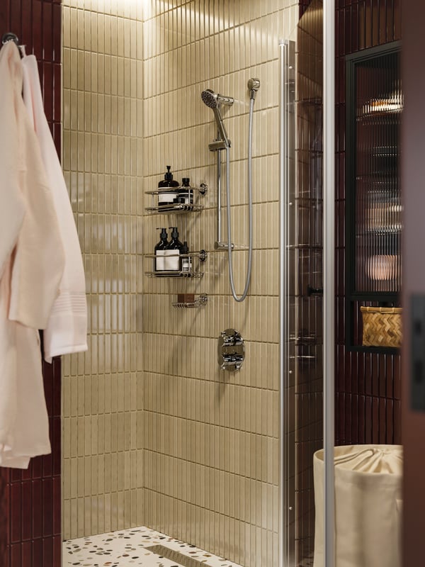 Une salle de bains avec un ensemble de douche VOXNAN avec mitigeur thermostatique, un carrelage beige et un rangement contenant des articles de toilette et une éponge.