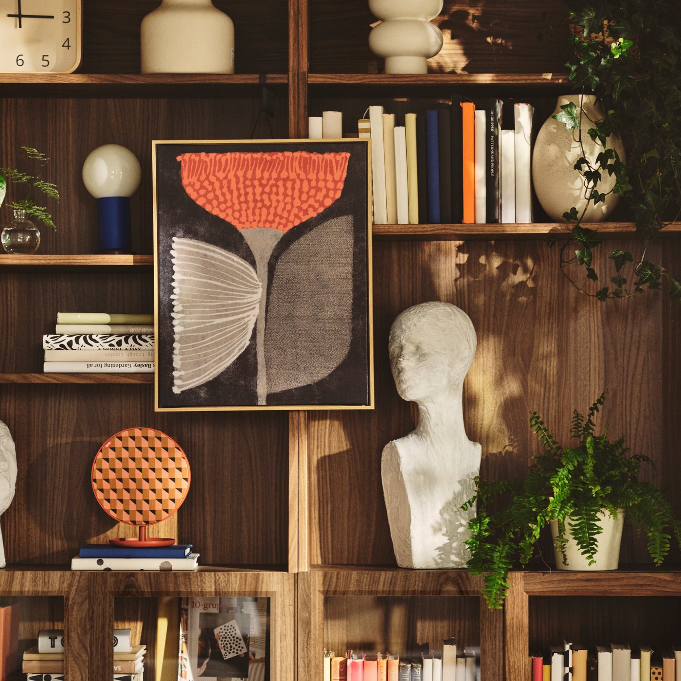 A living room features a BLODFLÄDER picture with an orange flower design on a wall above wooden shelves with decor.