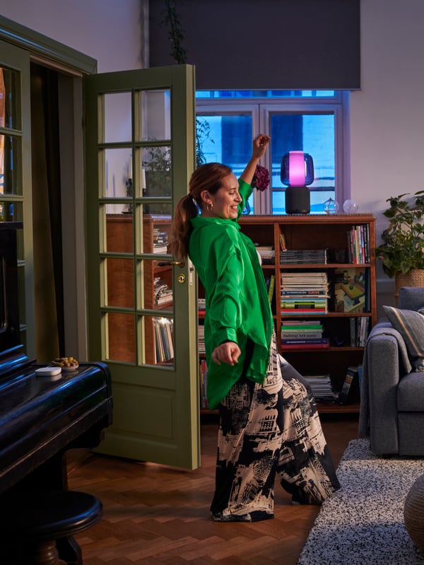 A lady in a green blouse is dancing in the living room. A smart white SYMFONISK table lamp is glowing with purple light.
