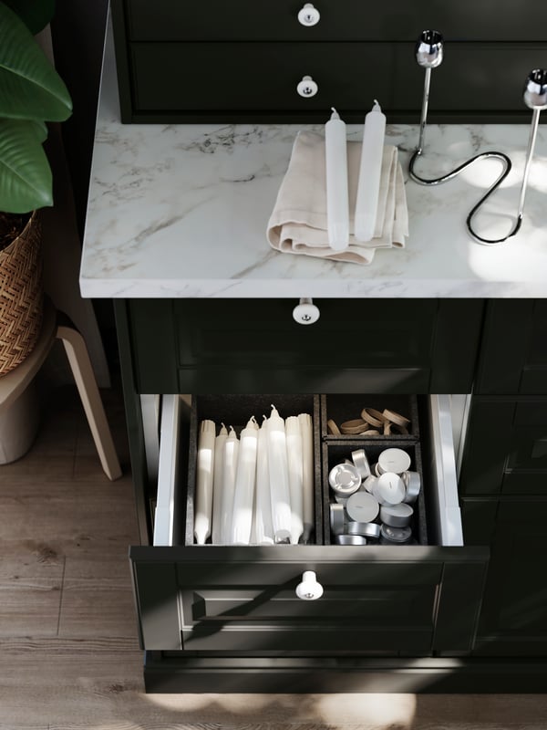 An open drawer in a cabinet with black BODBYN fronts shows interior organisers with dining room items sorted inside them.
