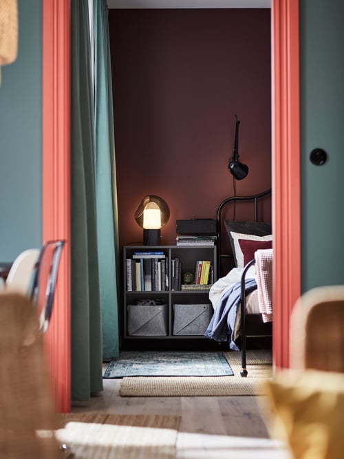 Blick durch ein Wohnzimmer in ein Schlafzimmer mit einem EKET Schrank mit 4 Fächern, u. a. mit einem SYMFONISK Regal-WiFi-Speaker, einer EVEDAL Tischleuchte und SANELA abdunkelnden Gardinenschals.
