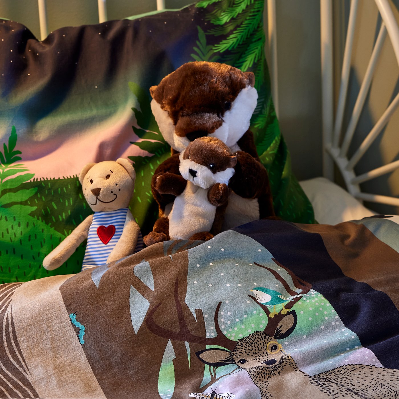 L'angolo di un letto per bambini con biancheria da letto SKOGSDUVA. Nel letto, con le coperte rimboccate, un peluche FABLER BJÖRN e un set di due peluche a forma di lontra.