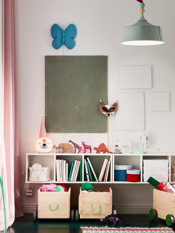 A white wall with SMÅGÖRA shelf units holding toys and books. Below them stand three FLISAT toy storage with wheels.