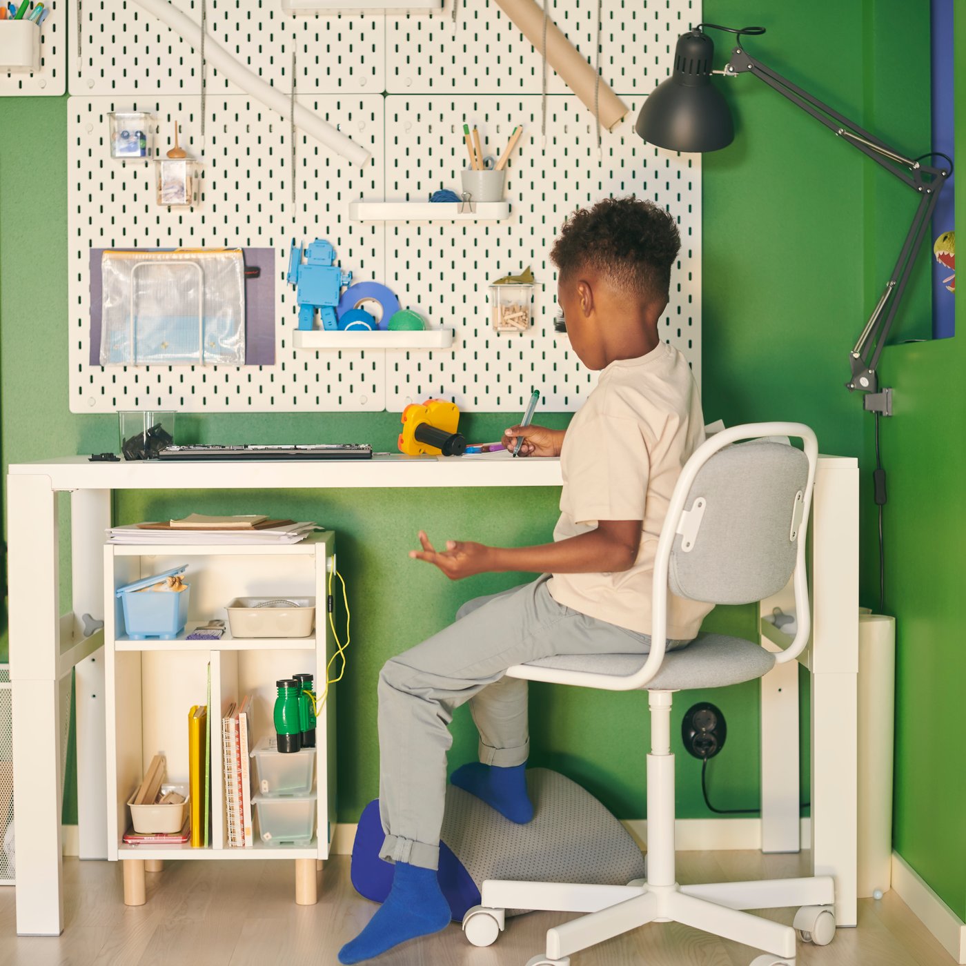 Ein Kind auf einem ÖRFJÄLL Kinderdrehstuhl an einem PÅHL Schreibtisch unter einer weißen SKÅDIS Lochplattenkombination an einer grünen Wand.