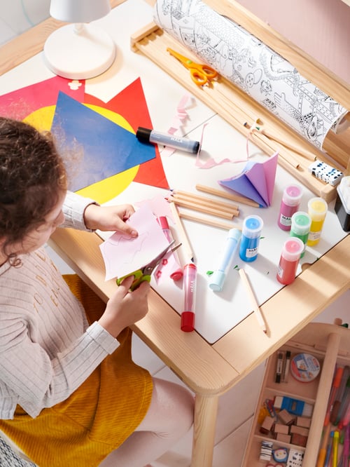 A child sitting at a FLISAT children’s table cutting some paper. MALA paints and a LUSTIG colouring roll are on the table.