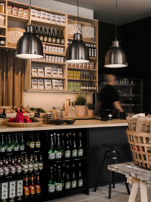 En bar-liknande disk i delikatessbutiken Casa Navarro med hyllor, vinflaskor, färska grönsaker på disken och kylda drycker under. Tre SVARTNORA pendellampor lyser upp rummet.