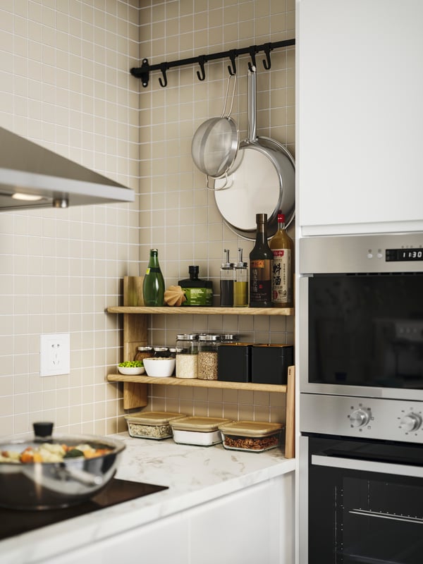 A kitchen with KLAVRESTRÖM wall shelves stocked with jars, oils, and pans hanging from hooks above the tiled backsplash.