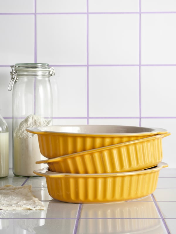 Three POETISK oven dishes in light yellow are stacked on a white tiled surface, next to some dough and in front of two jars.