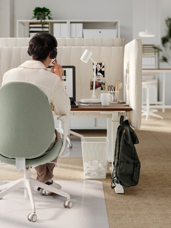 Ein Mann mit einem Headset sitzt in einem grünen GRÖNFJÄLL-Bürostuhl an einem nussbaumfarbenen/weißen Schreibtisch mit MITTZON-Akustikschirmen für den Schreibtisch.