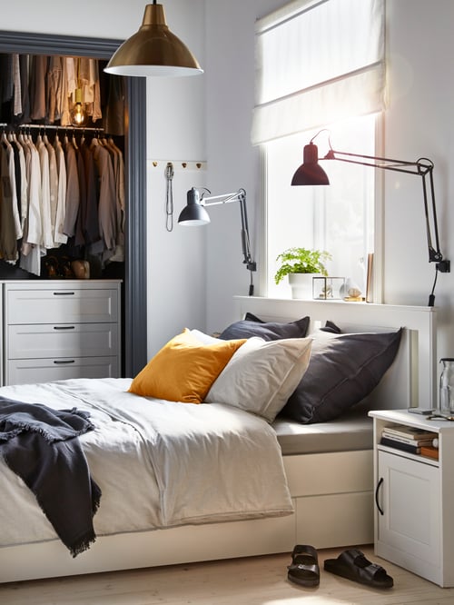 Une chambre avec une armoire ouverte et un lit SONGESAND blanc avec quatre boîtes de rangement, une literie grise et une table de chevet blanche.