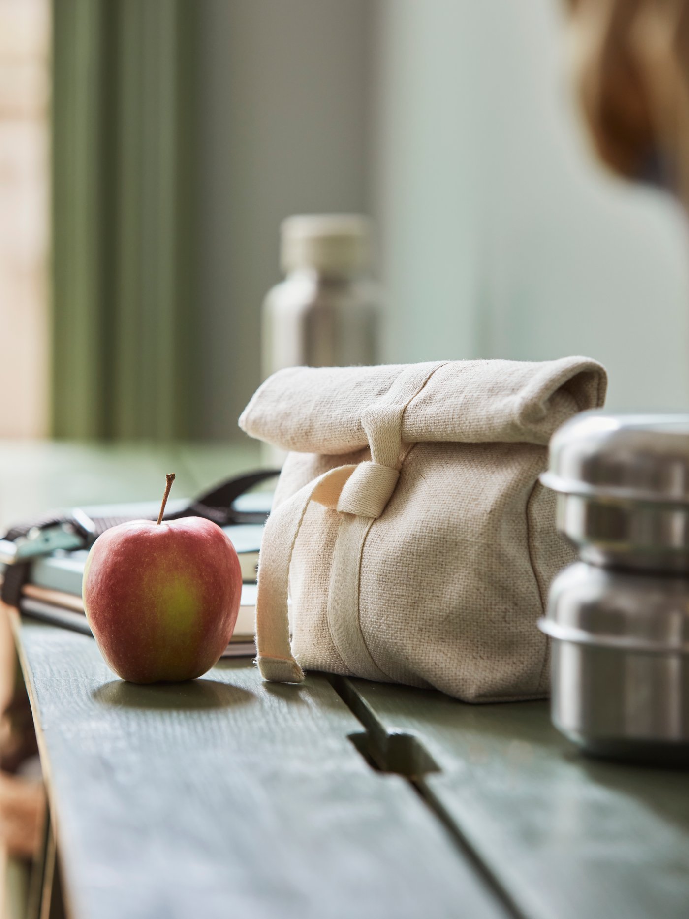 A closed HAJMAL snack bag in natural fabric, standing on a PERJOHAN bench beside an apple and a LÄTTUGGAD snack container.