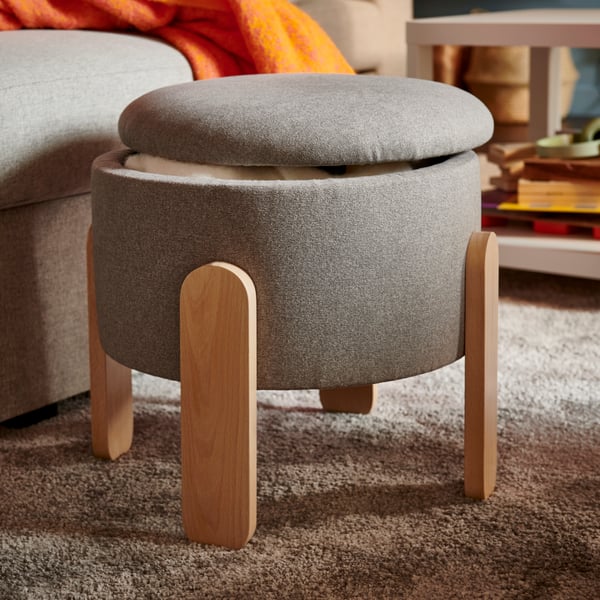 A grey FÖRLUNDA pouffe with a removable top, positioned beside a sofa, featuring soft fabric and wooden legs.