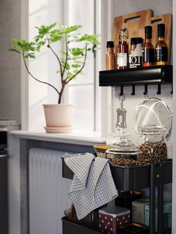 A RÅSKOG cart with jars, a tea towel, and various kitchen items, placed next to a window with a potted plant on the sill.