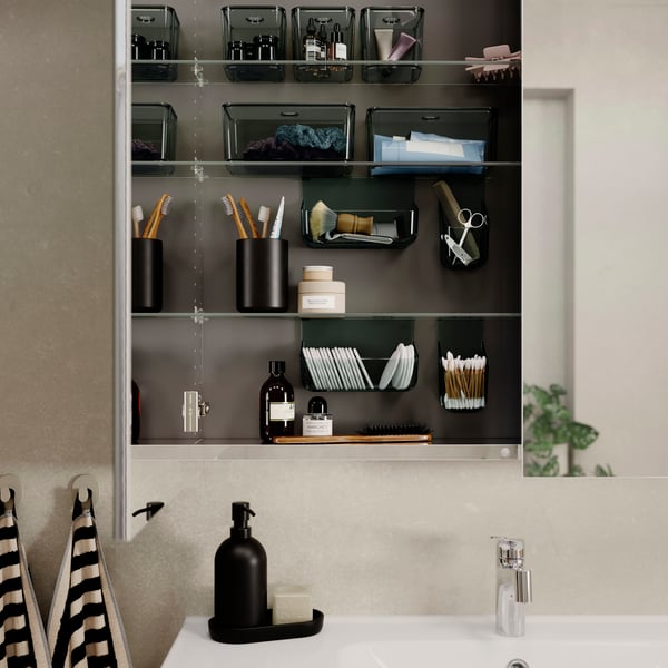 A bathroom with a high cabinet with open doors showcasing various VISSLAÅN inside organisers in transparent grey.