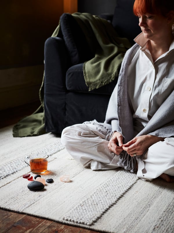A woman sits on a light-coloured rug in a living room, with crystals, stones and a glass mug in front of her.