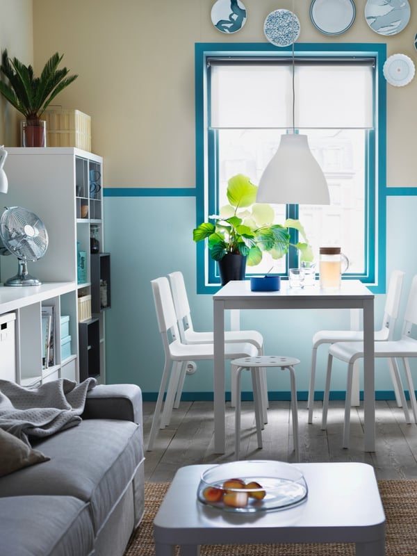 A white table with a mix of rattan and wooden chairs around it, and with crockery and glasses on top in a white dining room.