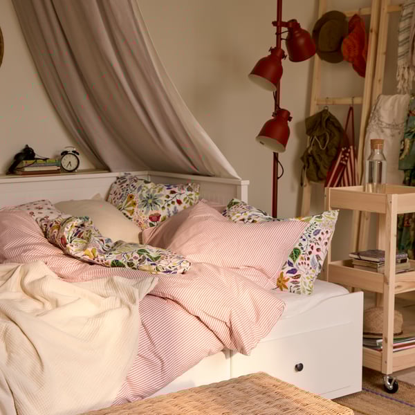 HEMNES day-bed with a mix of bedlinen; the striped SOLFIBBLA and the floral patterned BRUDKRUSBÄR. HEKTAR floor lamp in back.