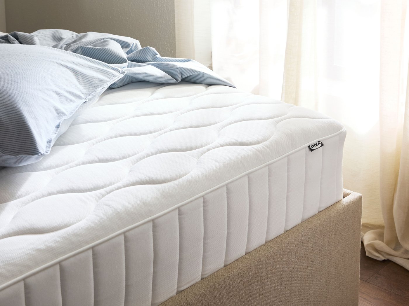 Close-up of thick white mattress in a beige bed frame. Window with light shining through in the back.