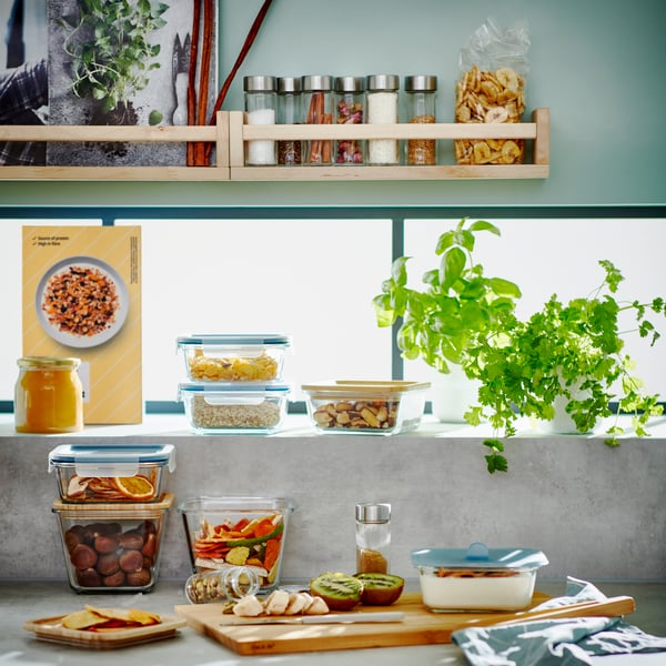 Plan de travail de cuisine avec un repas compartimenté dans une boîte repas, une planche à découper avec des tomates et des herbes aromatiques fraîchement coupées.