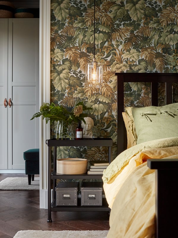 A KORNSJÖ side table used as a bedside table stands next to a HEMNES bed. It holds decorative items, books and boxes.