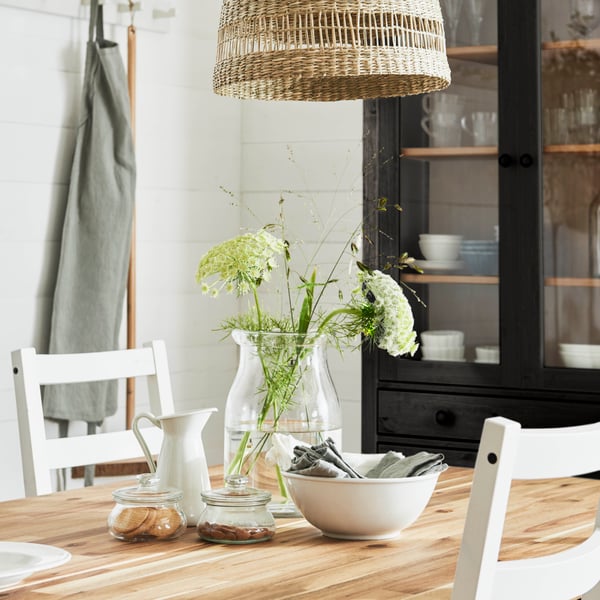 Ein gemütliches Esszimmer mit einem SKOGSTA Tisch aus Holz, auf dem eine Glasvase mit Blumen, eine weiße Schale und Gläser stehen. Darüber hängt eine Korblampe. Im Hintergrund steht ein schwarzer HEMNES Schrank.