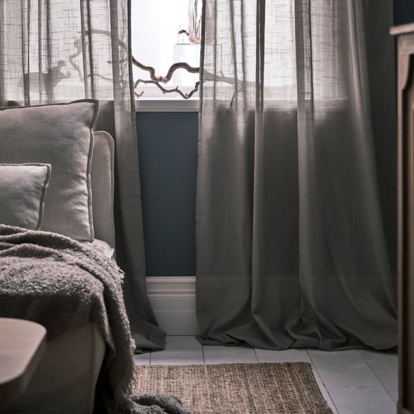 Tende sottili SILVERLÖNN beige drappeggiate sul pavimento davanti a una finestra accanto a un divano di colore naturale su un tappeto in iuta LOHALS.