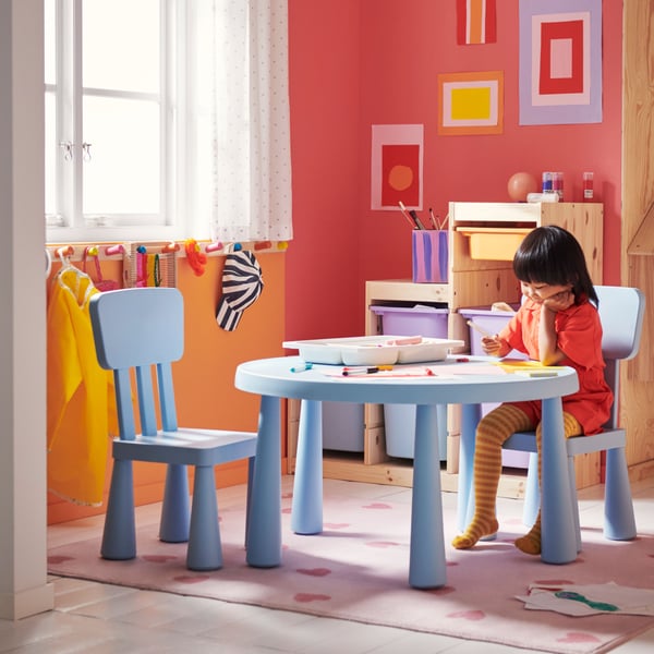 A child sitting on a MAMMUT chair at a MAMMUT children’s table drawing a picture. A tray of art materials is on the table.
