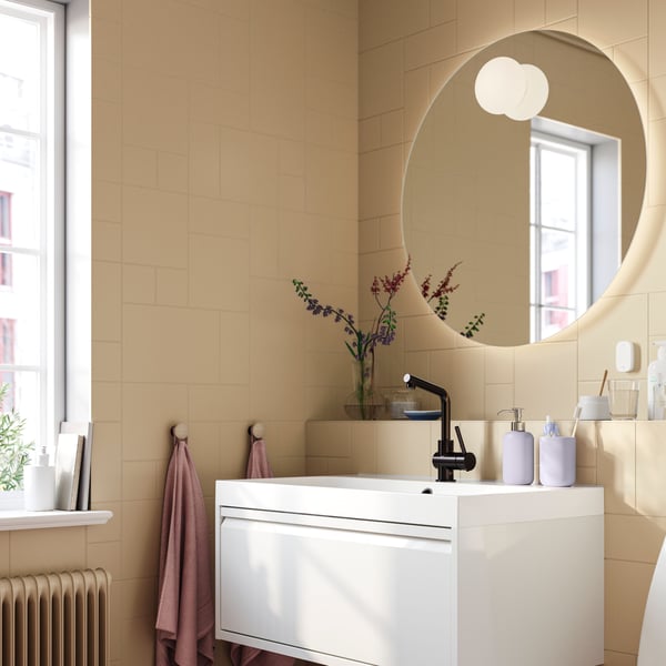 A circular FAXÄLVEN mirror with built-in lighting is backlit, on a recessed pale yellow tiled wall above a white wash-stand.