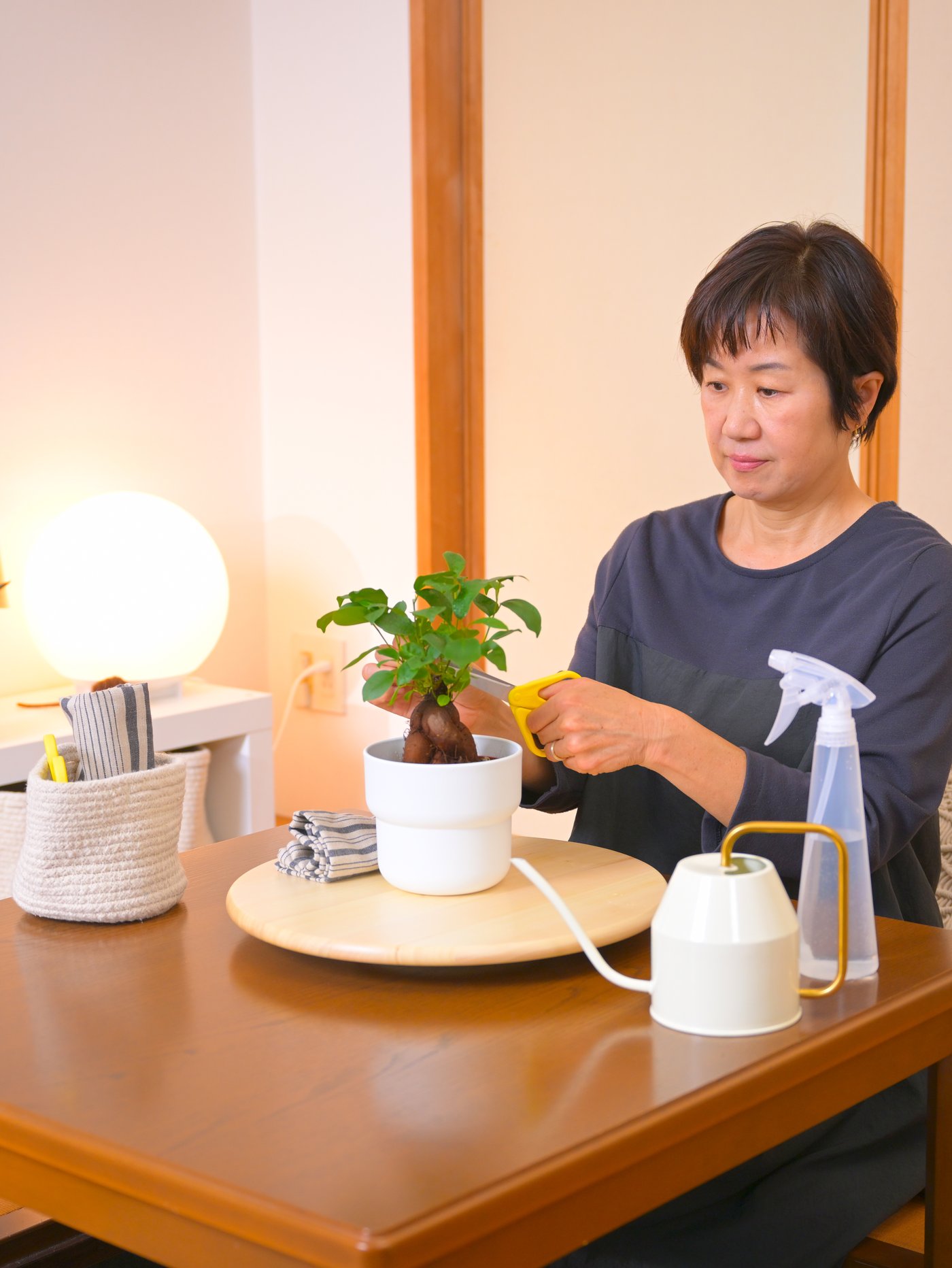 Eine Frau sitzt an einem Tisch und verziert eine Vase in der eine Pflanze ist. Auf der Seite steht eine Lampe die leuchtet.