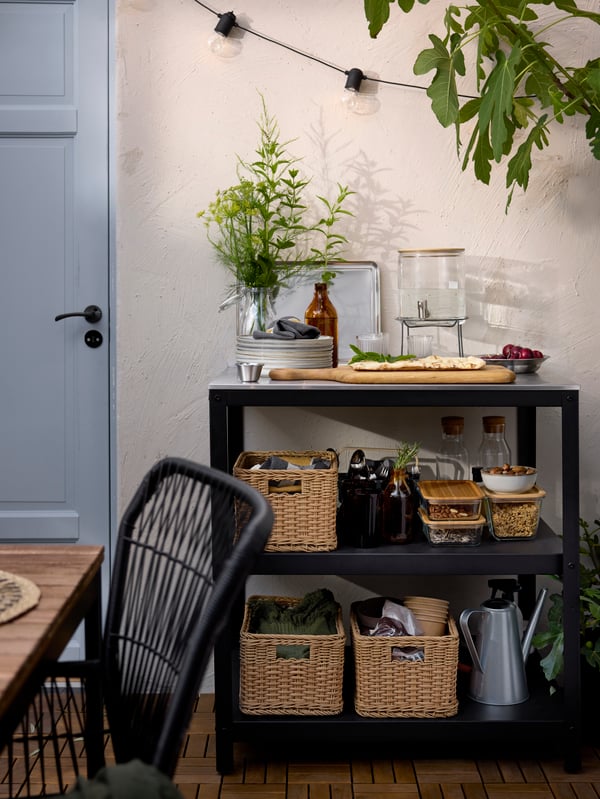 A patio with a GRILLSKÄR kitchen island in stainless steel with open shelves filled with food and dinnerware in baskets.
