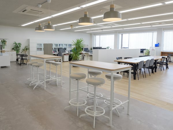 白いハイテーブルとスツールを中心に、奥にワークデスクと観葉植物が並ぶ開放的なオフィスラウンジ。