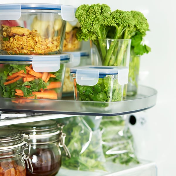 An open refrigerator stocked with various food items. Visible are clear containers with blue lids, one containing a mixed salad, another with what appears to be a grain-based dish, and a third holding cut vegetables including carrots and broccoli. There is also a glass jar filled with a red sauce or preserve. The focus on fresh and prepared foods suggests an emphasis on healthy eating or meal preparation.