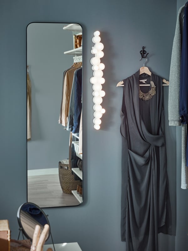 Un miroir sur pied sur un mur bleu-gris, un éclairage original, un crochet noir et une robe grise sur un cintre.