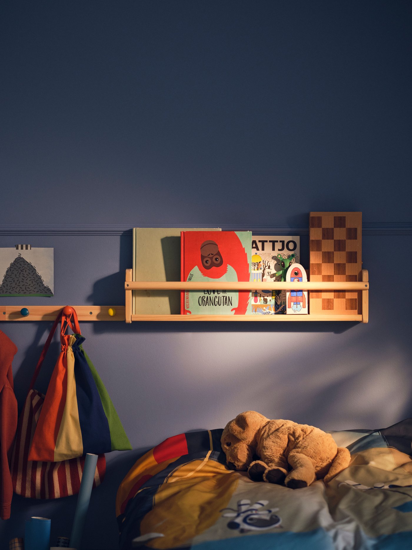 A light wood FLISAT wall storage holding various children’s storybooks on a blue wall beside a FLISAT knob rack with 4 knobs.