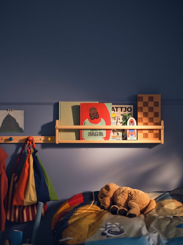 A light wood FLISAT wall storage holding various children’s storybooks on a blue wall beside a FLISAT knob rack with 4 knobs.