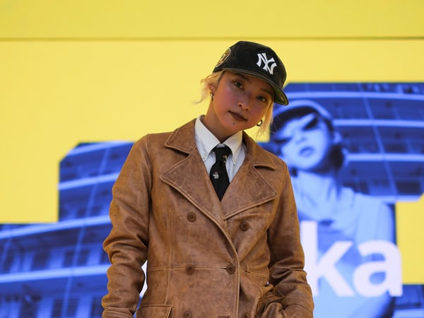 Kyoka standing on a stage with a leather coat, hat, tie and white shirt with a backdrop of her in the colour blue and yellow that comes from the digital display used on stage during Milan design week 2024 at the IKEA venue.