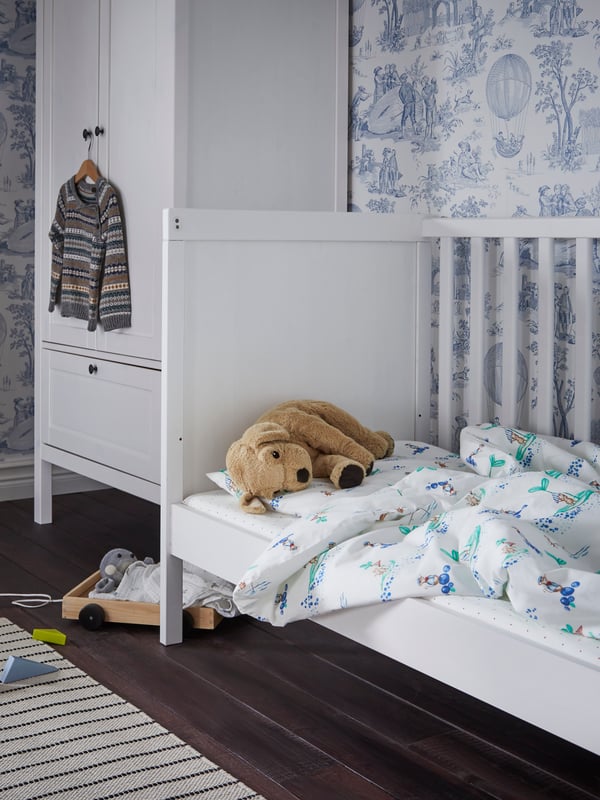 A white SUNDVIK cot with one side removed and RÖDHAKE bed linen stands beside a SUNDVIK wardrobe in a child’s bedroom.