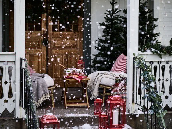 Juldekorerad veranda med snöfall, röda lyktor, två trästolar med filtar, ett bord med te och snacks samt en julgran vid en rustik trädörr.