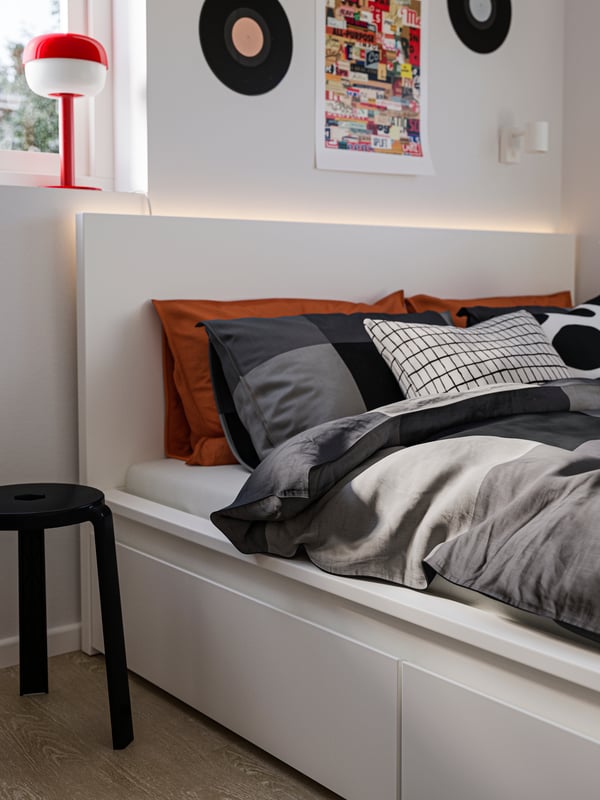 Close-up of a white MALM bed with drawers, BRUNKRISSLA bedding, a black stool by the side and a red lamp in the window.