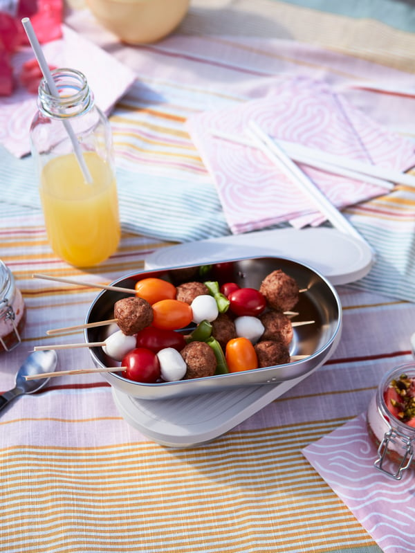 An opened food container holding skewers with HUVUDROLL plant balls, tomatoes and more placed on a striped cloth.