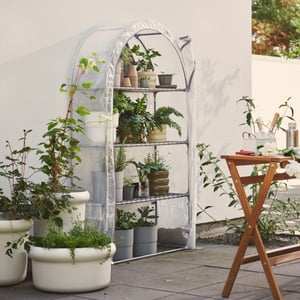 A garden with a transparent white ASPAREN greenhouse filled with plants on shelves next to a wooden table and green plants.