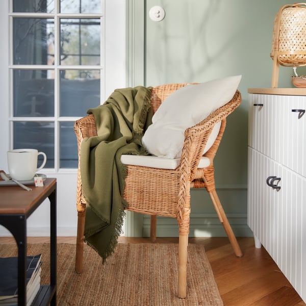 Ein gemütlicher Sessel aus Rattan, bedeckt mit einem weichen Kissen und einem grünen Überwurf. Ein weißer Schrank und eine Tasse auf dem Tisch ergänzen den Raum, der durch das natürliche Licht, das durch das Fenster fällt, eine einladende Atmosphäre erhält.