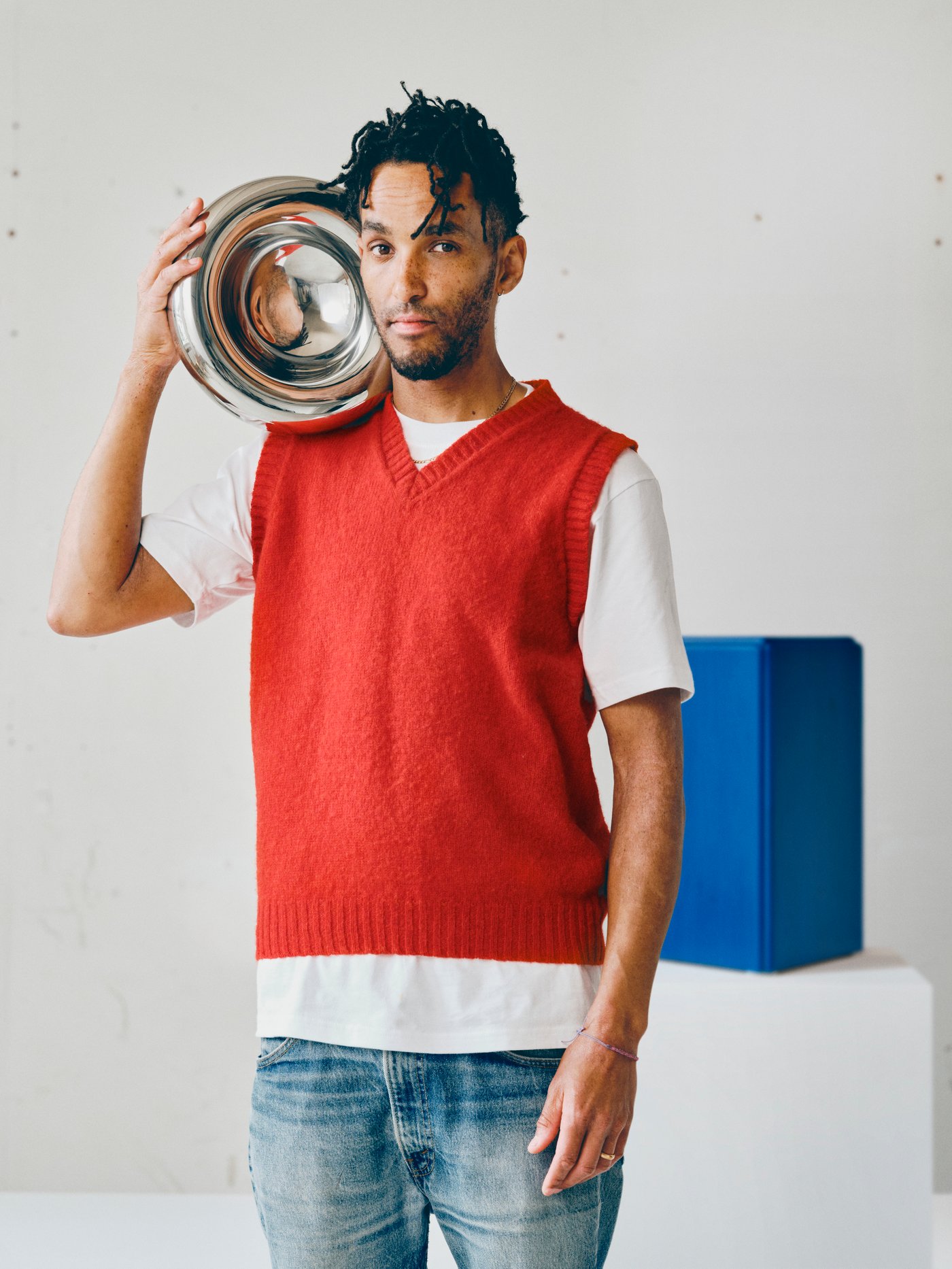 Andu Masebo holding a metallic bowl close to his head, behind him there’s one of his blue sculptures.