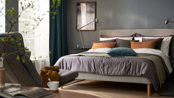 Bedroom with dark grey walls and a bed with white-grey-orange bedding, an armchair in foreground and windows in back.