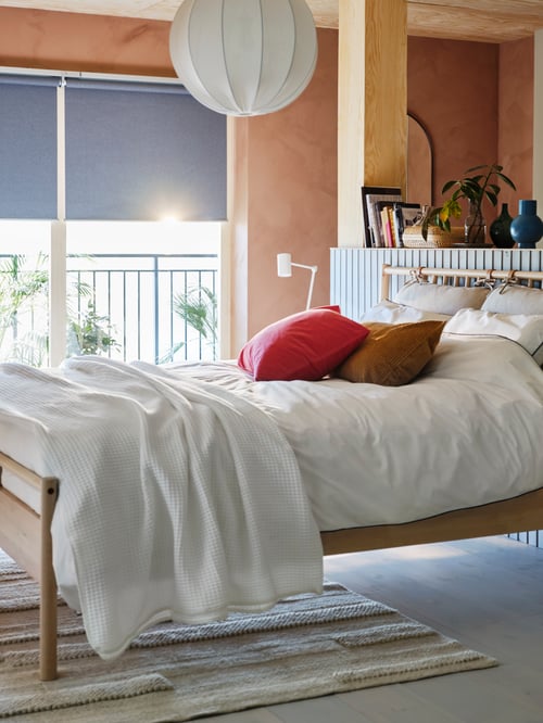 A modern bedroom featuring a bed frame, a white bedspread, a block-out roller blind, a pendant lamp shade, and a duvet cover.