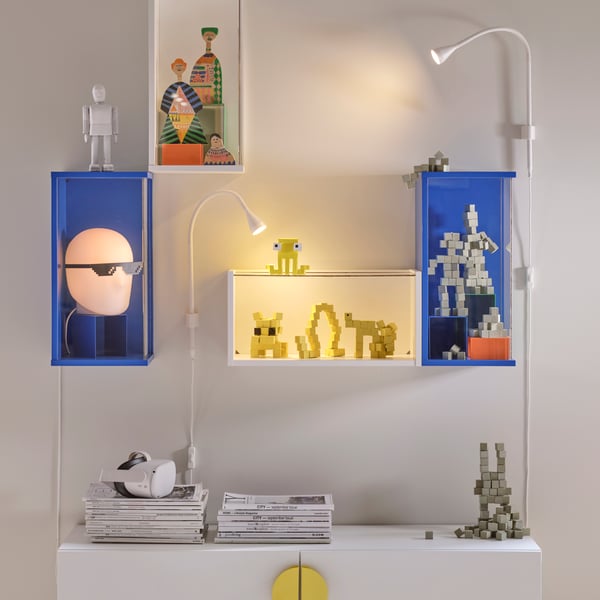 A white wall with several display cabinets containing gaming accessories, lit by white NÄVLINGE LED wall/clamp spotlights in the living room.