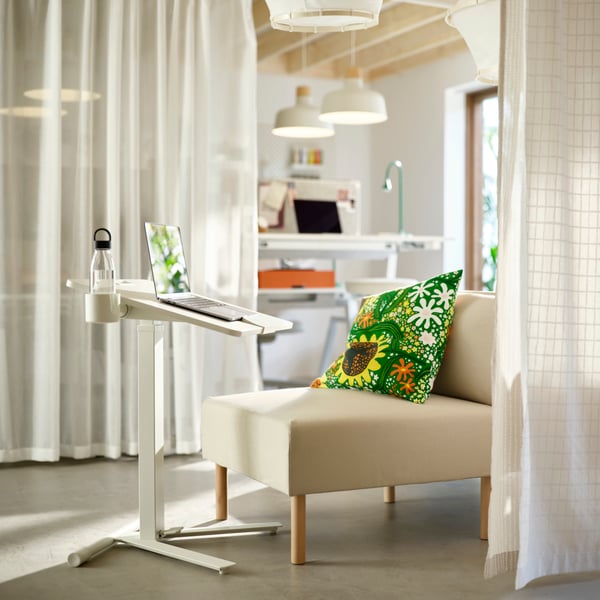 Un canapé blanc simple avec un support de bureau tenant un ordinateur portable. Espace de travail commun en arrière-plan ; rideaux de séparation de chaque côté.