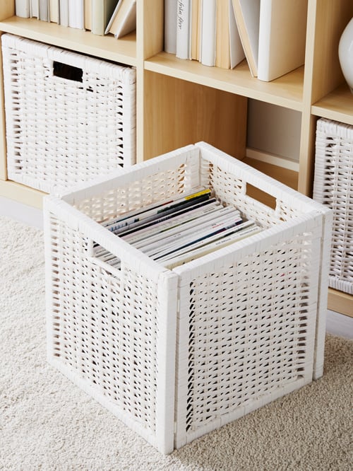 A white woven, cubic BRANÄS basket holds magazines while standing on the floor. Behind it is a shelf with matching baskets.